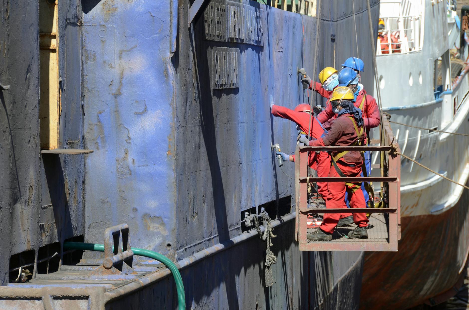 men on a hanging platform repairing the outside of a ship in a shipyard